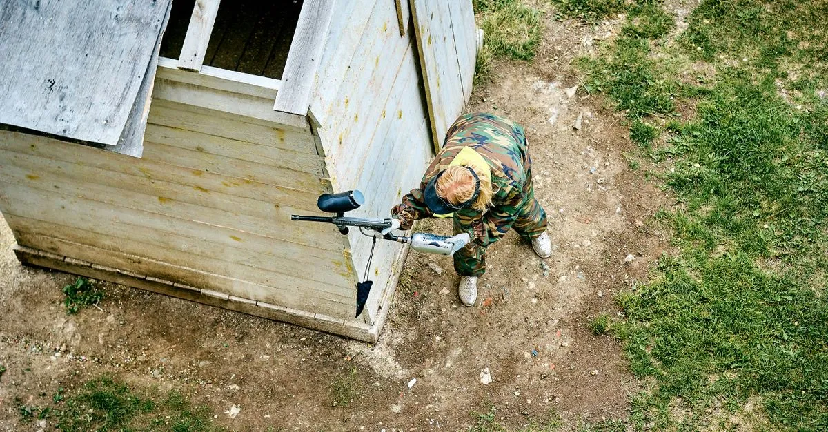 Camouflaged player holding a concealed position beside a hut with a paintball sniper rifle setup